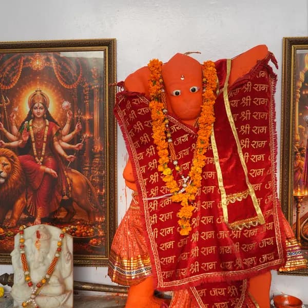 Hanuman ji on the altar in saffron sindoor, wrapped in a red velvet cloth printed with Jai Shri Ram and hung with marigold garlands, Durga in a gold frame and a small marble Ganesha to the left.