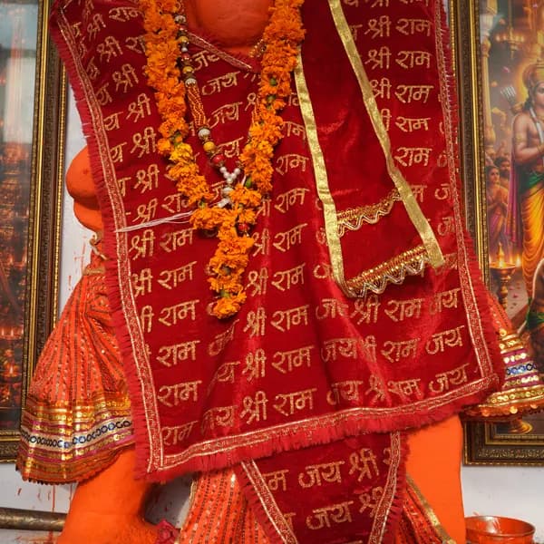 Close view of Hanuman ji's torso, red velvet shawl with gold Shri Ram lettering, stacked marigold malas, and Rama with a bow at the edge of a neighbouring frame.
