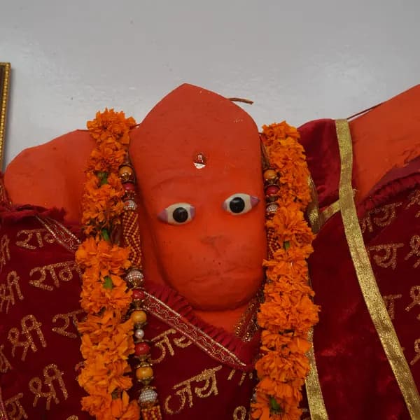 Face of the saffron Hanuman ji murti, large painted eyes and a silver mark on the forehead, marigold garlands at the neck over a red cloth with gold Jai Shri Ram script.
