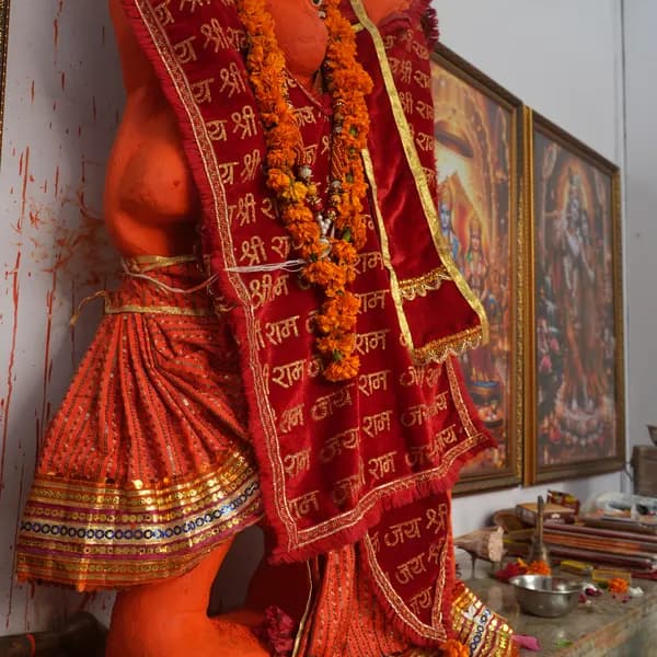 Hanuman ji from the shoulder, red velvet shawl over a sequined saffron dhoti, a marigold mala, incense and bowls on the marble ledge, Ram Darbar and Radha Krishna in gold frames.