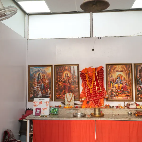 Shrine room looking toward the altar, Hanuman ji under a hanging brass bell, gold-framed deities on the white wall, a white hundi with a QR code on the red-draped ledge.