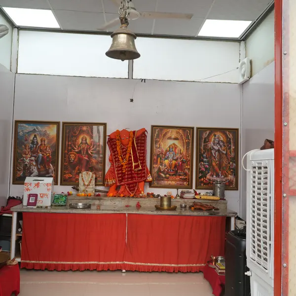 Wide interior of the shrine, Hanuman ji on the red-draped marble altar, four gold-framed pictures, a brass bell and ceiling fan, an air cooler against the right wall.