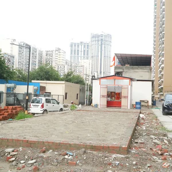 Construction plot in front of the small white shrine, a brick-lined rectangle of bare earth, Noida apartment towers behind, a saffron flag on the shrine roof.