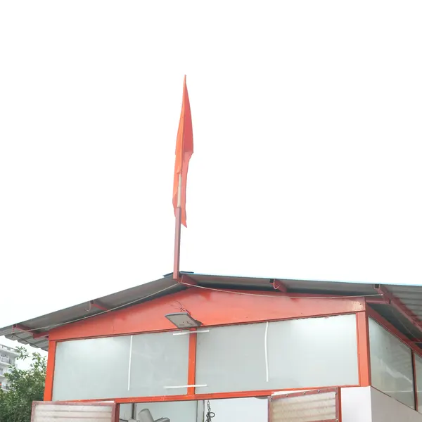 Saffron flag on the shrine roof against a bright sky, white and orange gable, a floodlight under the peak.