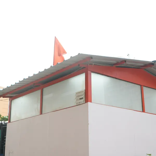 Low corner of the white shrine, red metal frame and corrugated roof, saffron flag against an overcast sky.