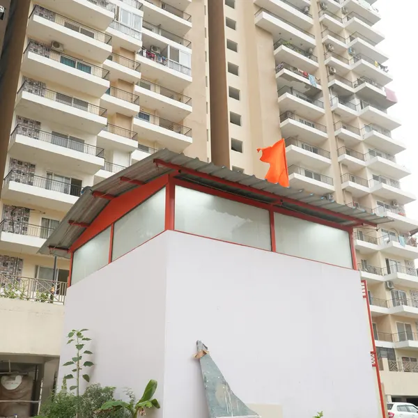 White shrine with red-trimmed roof and a saffron flag, a tall beige apartment block filling the background.