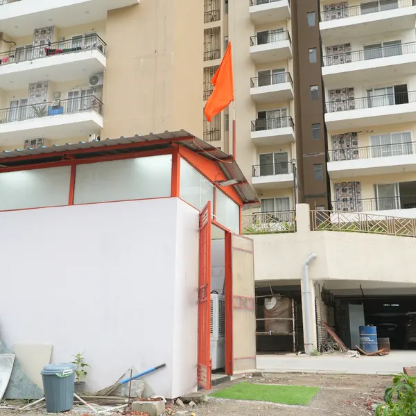 Side of the shrine with orange metal doors open, a patch of green turf at the entrance, saffron flag flying, apartment towers and water tanks behind.