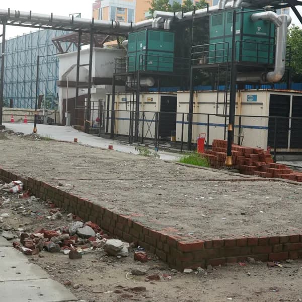 Bare rectangular plot edged with new red bricks, stacked bricks to the right, generators on a steel platform, the shrine and Noida towers in the distance.