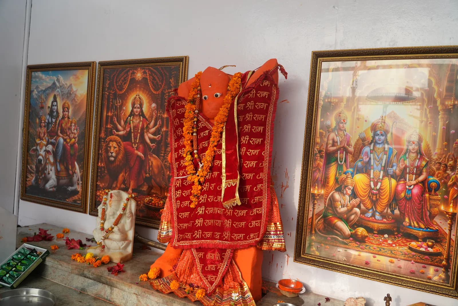 Garlanded Hanuman murti draped in red and saffron cloth, flanked by framed pictures of the deities